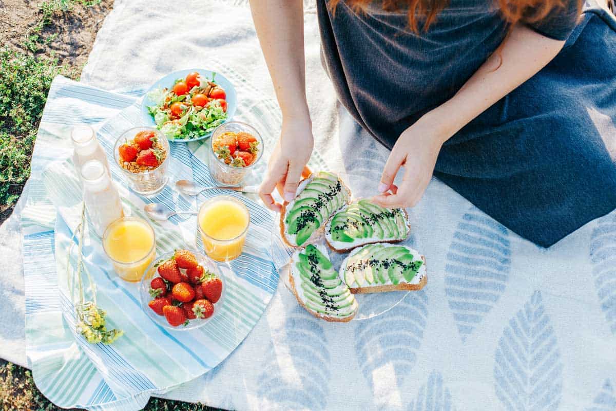 A person sitting on a blanket prepares avocado toast next to a picnic spread that includes salads, strawberries, orange juice, and glass bottles.