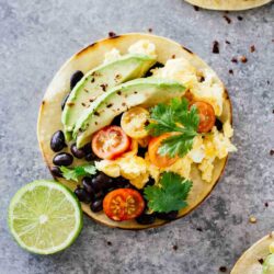 A tortilla topped with scrambled eggs, sliced avocado, black beans, cherry tomatoes, and cilantro. A lime half is placed next to it on a gray surface.