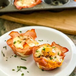 Two bacon-wrapped egg muffins garnished with chopped herbs on a white plate, with a muffin tray in the background.