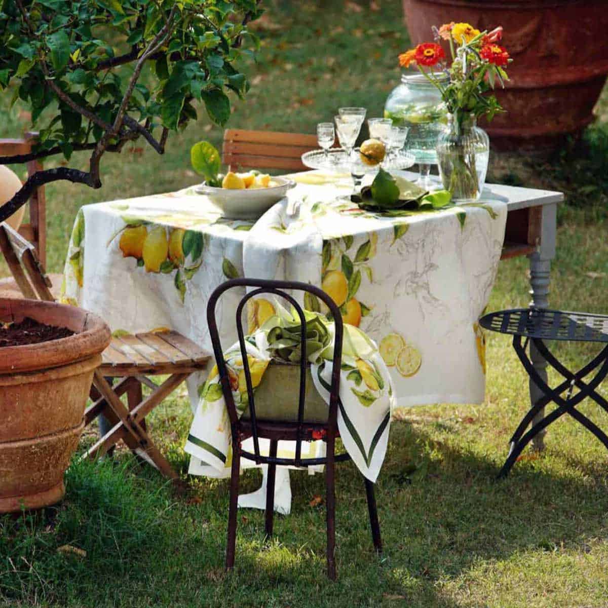 An outdoor table with a lemon-themed tablecloth, glassware, and a vase of flowers is set for dining. A chair draped with a matching cloth is in the foreground, surrounded by garden greenery.