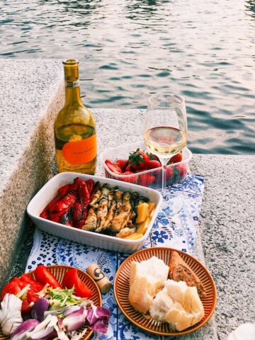 A picnic by the water in Italy with white wine, grilled vegetables, a salad, strawberries, and a plate with bread and a meat dish on a stone surface.