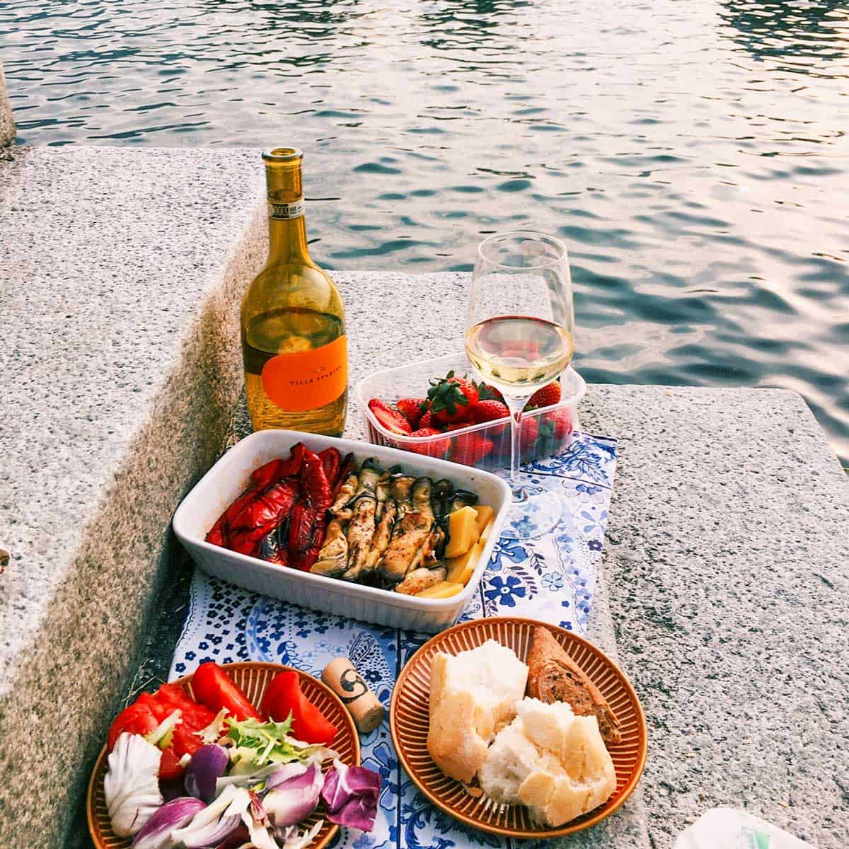 A picnic by the water in Italy with white wine, grilled vegetables, a salad, strawberries, and a plate with bread and a meat dish on a stone surface.