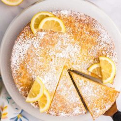 A round lemon cake dusted with powdered sugar, decorated with lemon slices. One slice is cut and partially removed from the cake.