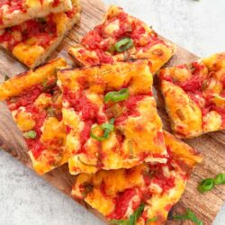 Close-up of rectangular focaccia slices with tomato sauce and green onion toppings, arranged on a wooden board.