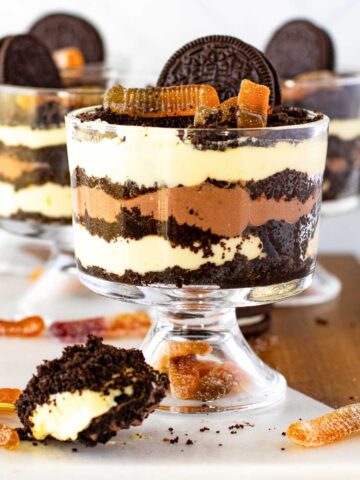 Glass bowls filled with layered Oreo Dirt Cakes with gummy worms. A spoonful of the dessert is placed in the foreground.