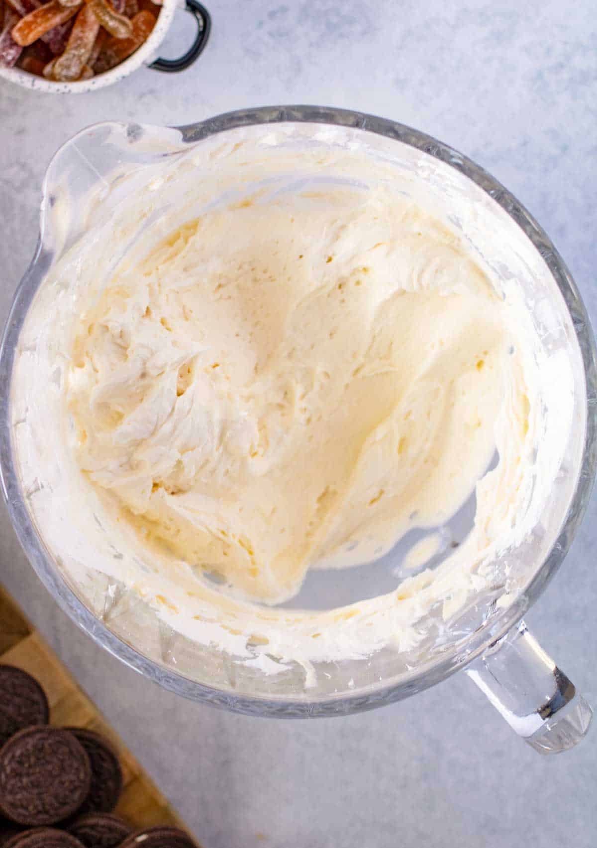 A glass bowl filled with whipped cream on a light gray surface, with a few pieces of chocolate cookies visible in the foreground.