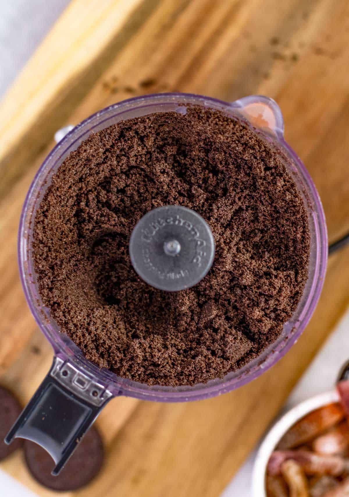 A food processor containing finely ground chocolate cookie crumbs on a wooden surface.