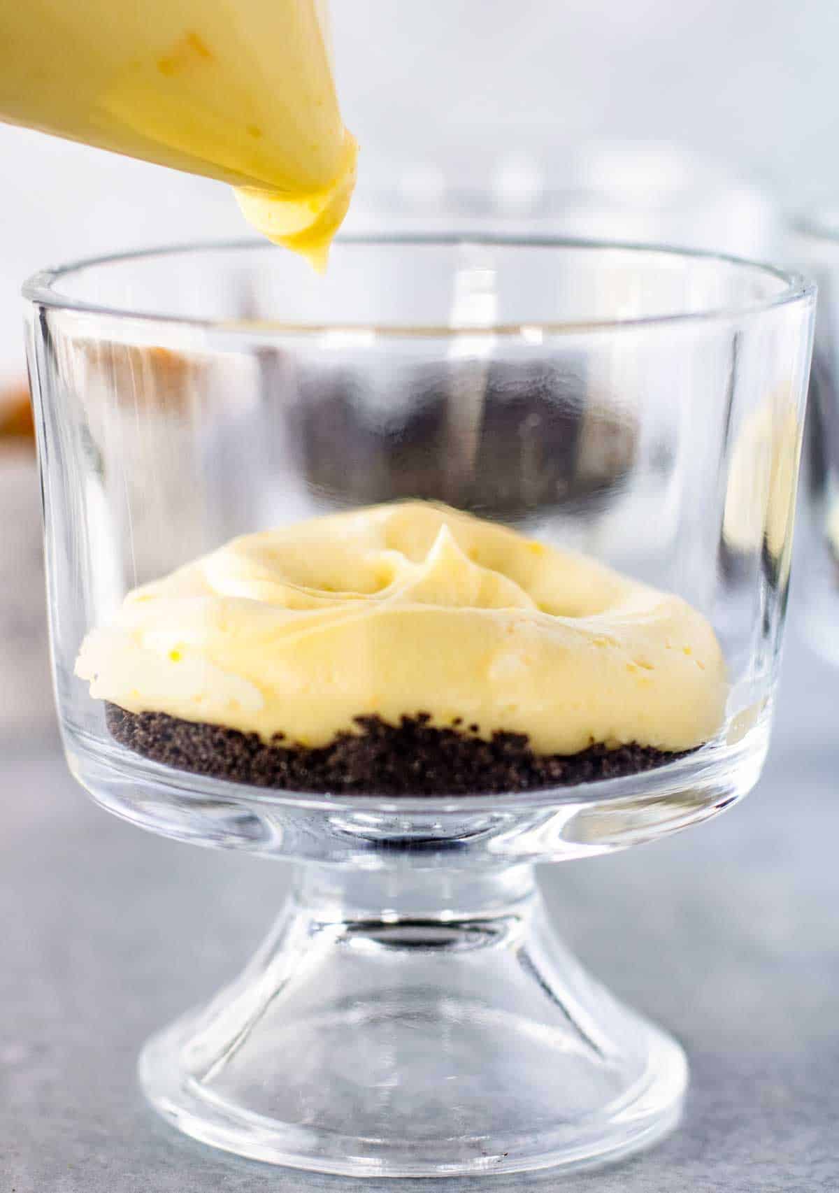 A glass cup with a layer of crushed cookie crumbs at the bottom and yellow cream being piped on top.