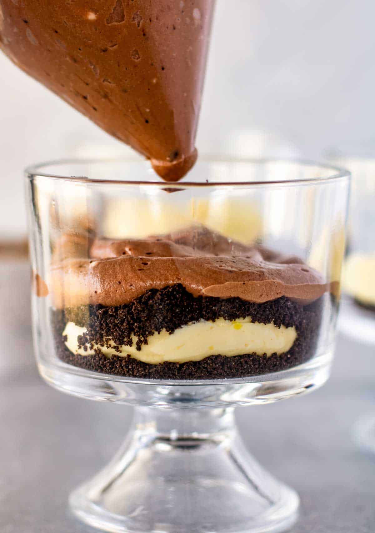 A glass dessert cup being filled with layers of chocolate mousse, crushed cookies, and cream.