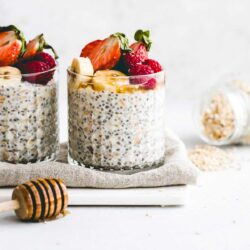 Two glass jars filled with chia seed pudding topped with sliced strawberries, bananas, and raspberries. A honey dipper and a jar of oats are nearby on a light surface.