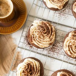 A brown coffee cup on a matching saucer next to a cooling rack with frosted Tiramisu Cookies dusted with cocoa powder.