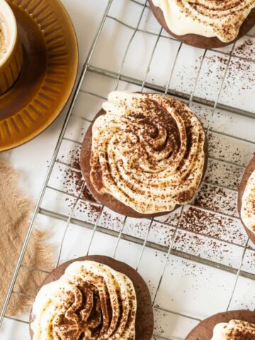 A brown coffee cup on a matching saucer next to a cooling rack with frosted Tiramisu Cookies dusted with cocoa powder.