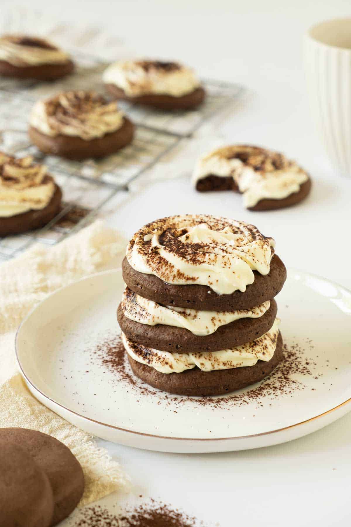 A stack of three Tiramisu Cookies with cocoa dusting on a white plate. In the background, more cookies are on a cooling rack.