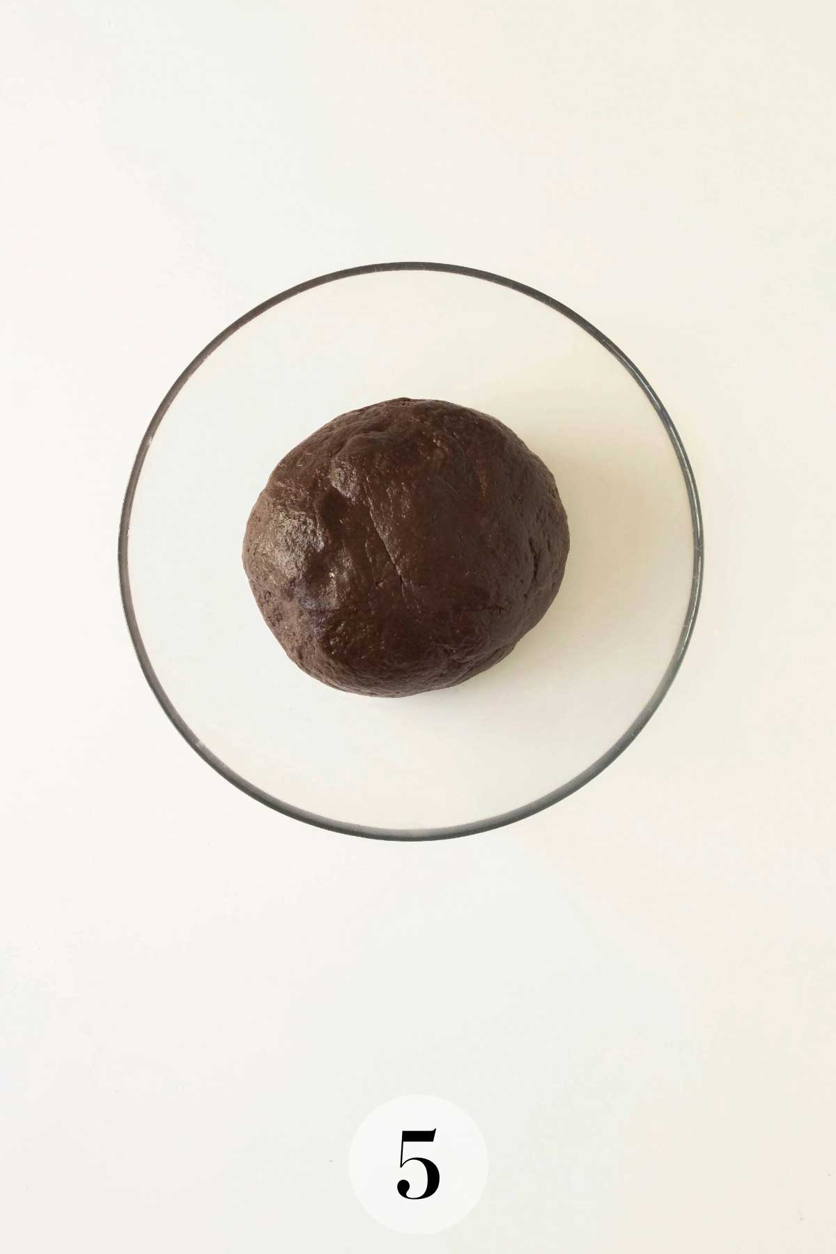 A ball of dark cookie dough placed in a clear glass bowl, shown against a plain white background.