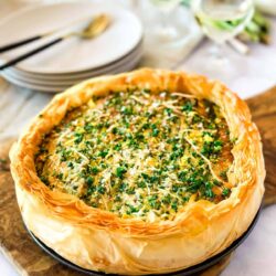 A savory pie with a golden-brown crust is garnished with chopped herbs. Plates, napkins, and glasses are set in the background, suggesting preparation for a meal.