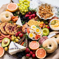 A colorful assortment of breakfast items, including bagels, fruits, cheeses, waffles, avocado, boiled eggs, and various spreads arranged on a surface.