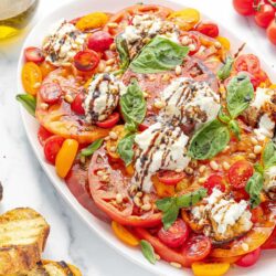 A colorful Caprese salad with sliced tomatoes, cherry tomatoes, basil leaves, pine nuts, and dollops of burrata cheese, drizzled with balsamic glaze, served on a white plate.