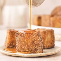 Close-up of three pieces of fried bread with a cinnamon sugar coating on a plate, being drizzled with syrup.