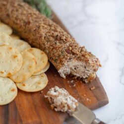 A log of cheese coated with chopped nuts sits on a wooden board beside round crackers, with a knife displaying a portion of the cheese.