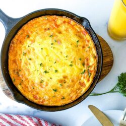 A baked frittata garnished with herbs in a cast iron skillet, placed on a wooden board next to a glass of orange juice and a parsley bunch.