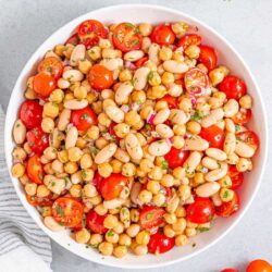 A bowl of mixed bean salad featuring chickpeas, white beans, halved cherry tomatoes, red onions, and chopped herbs.