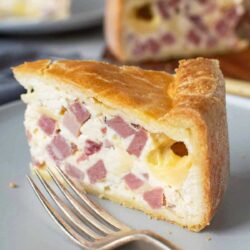 A slice of savory pie with ham cubes and cheese is served on a gray plate, accompanied by a silver fork.