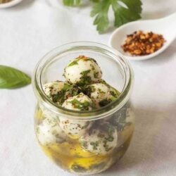 A jar filled with marinated mozzarella balls in olive oil, garnished with herbs. A spoon containing crushed red pepper flakes is visible in the background.