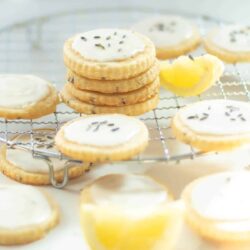 Stacked and individually placed frosted cookies on a cooling rack, with lemon slices and a few scattered lavender flowers around them.