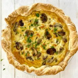 A baked quiche with a golden-brown crust, filled with mushrooms, cheese, and herbs, displayed on a white wooden surface.