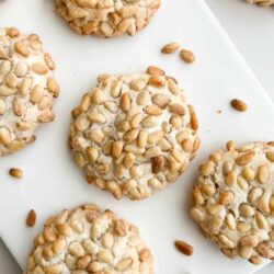 Five pine nut-topped cookies are arranged on a white surface, with scattered pine nuts around them.