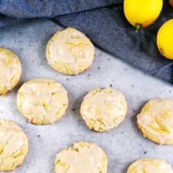 Eight glazed lemon cookies are laid out on a gray surface, with a blue cloth and whole lemons visible in the background.