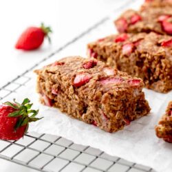 Close-up of strawberry oatmeal bars on a cooling rack with parchment paper. Fresh strawberries are seen in the background and foreground.