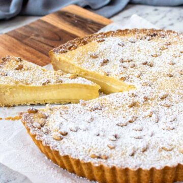 A sliced custard tart with powdered sugar on top, resting on parchment paper atop a wooden cutting board. One slice is being served.