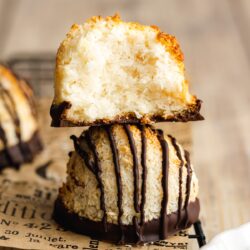 A partially eaten coconut macaroon with a chocolate base is stacked on top of another chocolate-drizzled macaroon on a newspaper-themed surface.