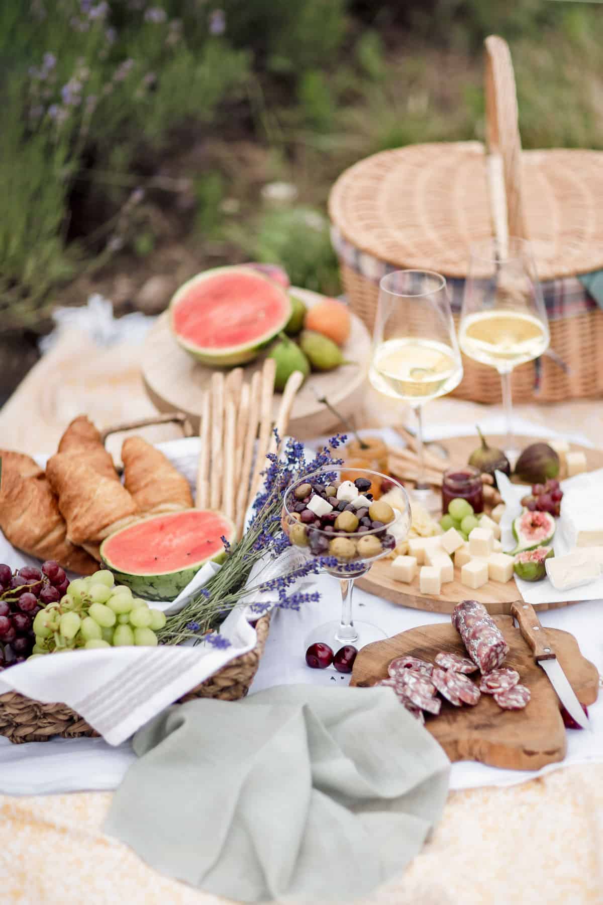 A picnic spread with wine glasses, croissants, watermelon slices, breadsticks, grapes, figs, cheese, salami, and a wicker basket on a blanket.