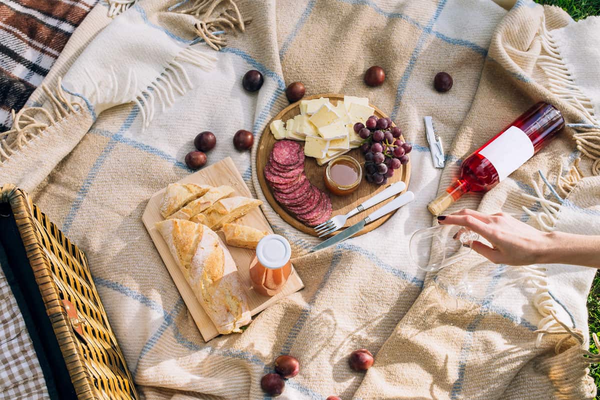 A picnic setup with sliced bread, cheese, salami, grapes, a bottle of rosé wine, and a person's hand reaching for a glass on a plaid blanket.