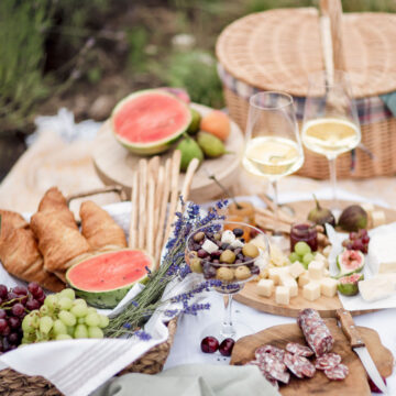 A picnic setup with croissants, watermelon, grapes, olives, cheese, salami, and two glasses of white wine on a table. A wicker basket is in the background.