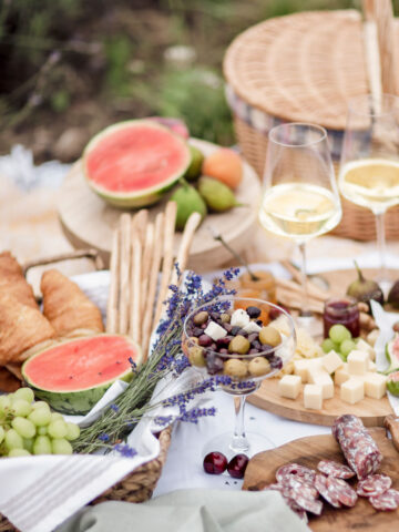 A picnic setup with croissants, watermelon, grapes, olives, cheese, salami, and two glasses of white wine on a table. A wicker basket is in the background.