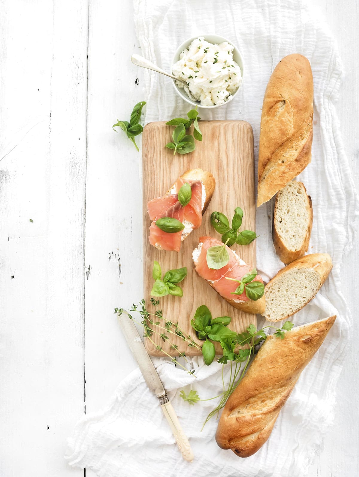 A wooden board with salmon on bread, garnished with herbs. Surrounding items include a bowl of cream cheese, two baguettes, slices of bread, herbs, and a knife on a white cloth.